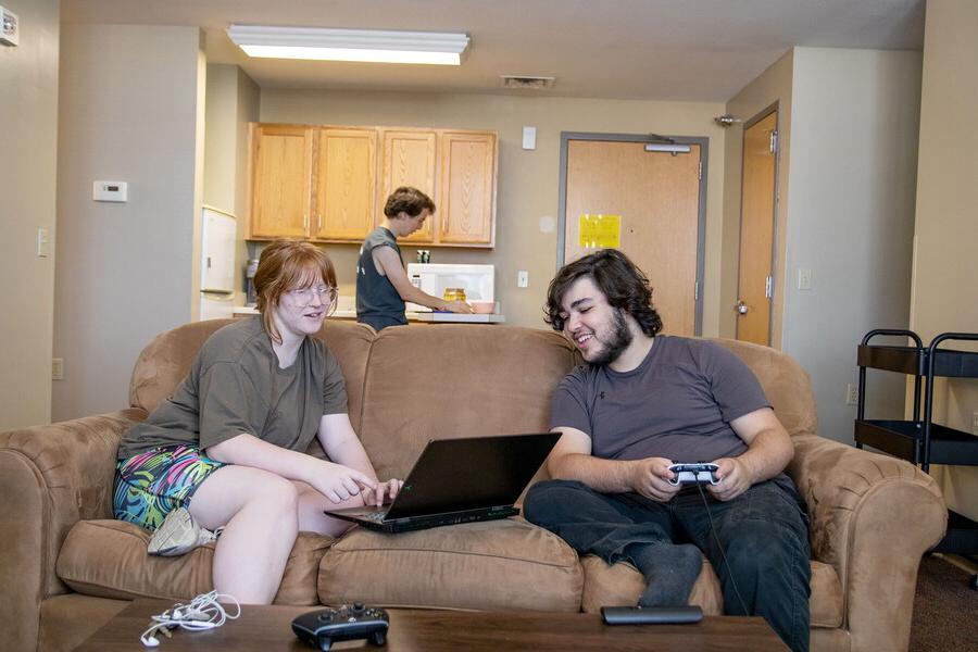 A male and female on a couch looking at a computer with a young man in the kitchen behind them.